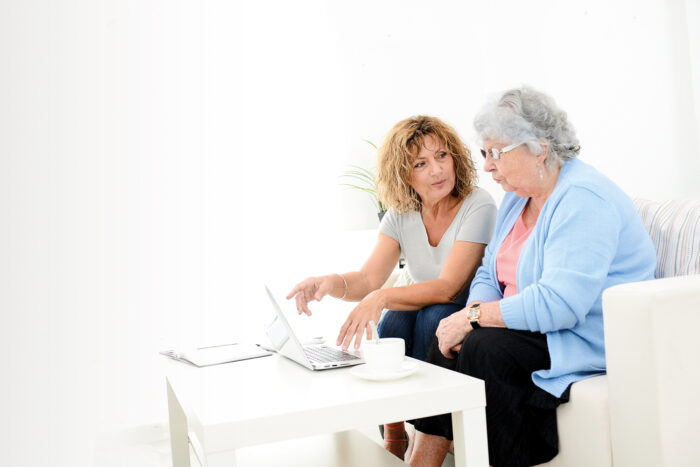 An elder woman sitting on a couch, with a younger woman who is pointing at a laptop that is on top of a coffee table, likely discussing Lasting Power of Attorney (LPA)